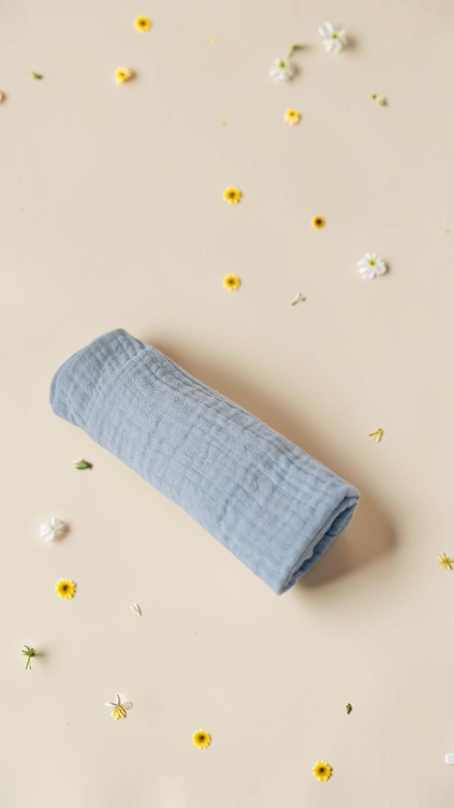 Cloudy Cotton blue organic muslin swaddle blanket rolled on a beige background with small scattered flowers.
